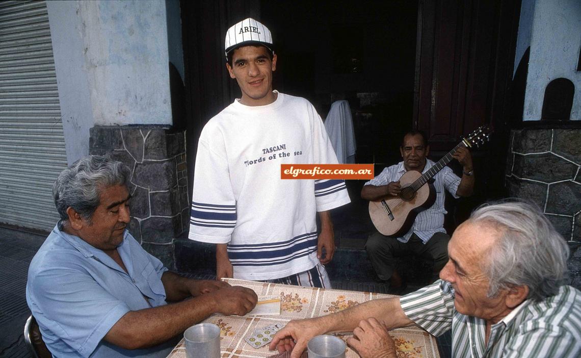 Imagen A Orteguita, feliz entre dos vecinos que juegan al truco y uno que toca la guitarra. 