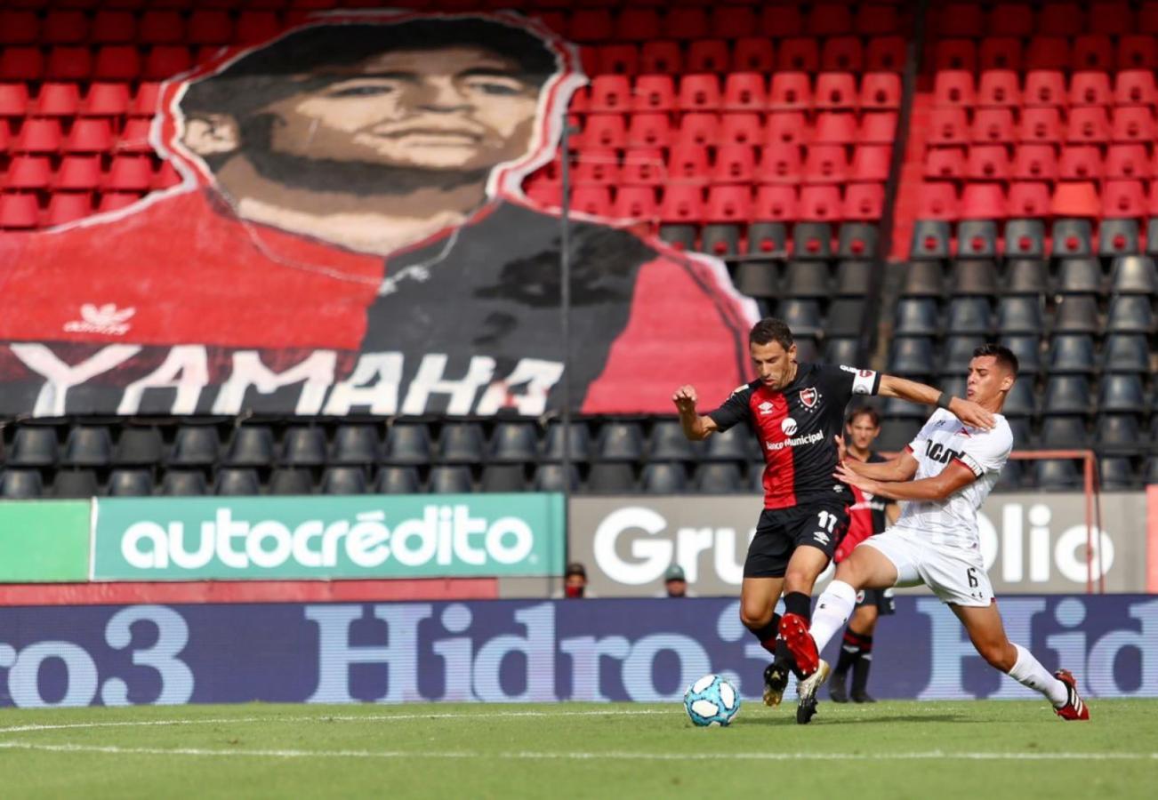 Imagen Diego eterno. Maxi Rodríguez domina la pelota ante Nazareno Colombo. De fondo, una bandera con la imagen de Diego y la casaca de Newell's, como observando el partido desde la platea.