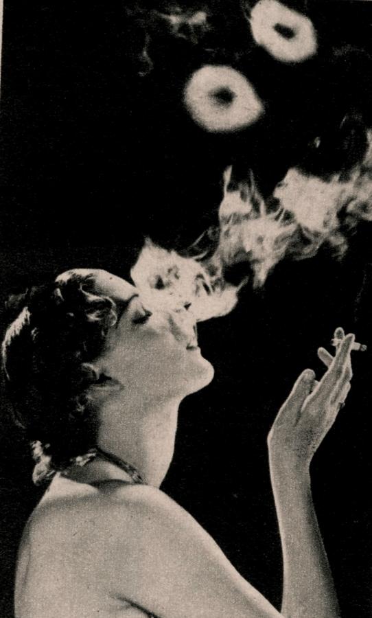 Imagen El humo se torna admirablemente dócil en los labios de esta hermosa mujer que oculta a medias su delicado rostro tras de una aureola luminosa que se difama.